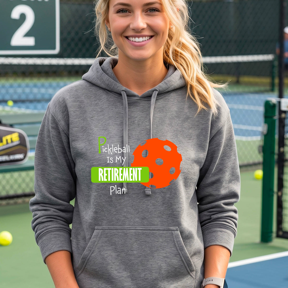 A woman smiling on a pickleball court wearing a gray hoodie with a graphic that reads 'Pickleball Is My Retirement Plan' alongside an orange pickleball illustration.