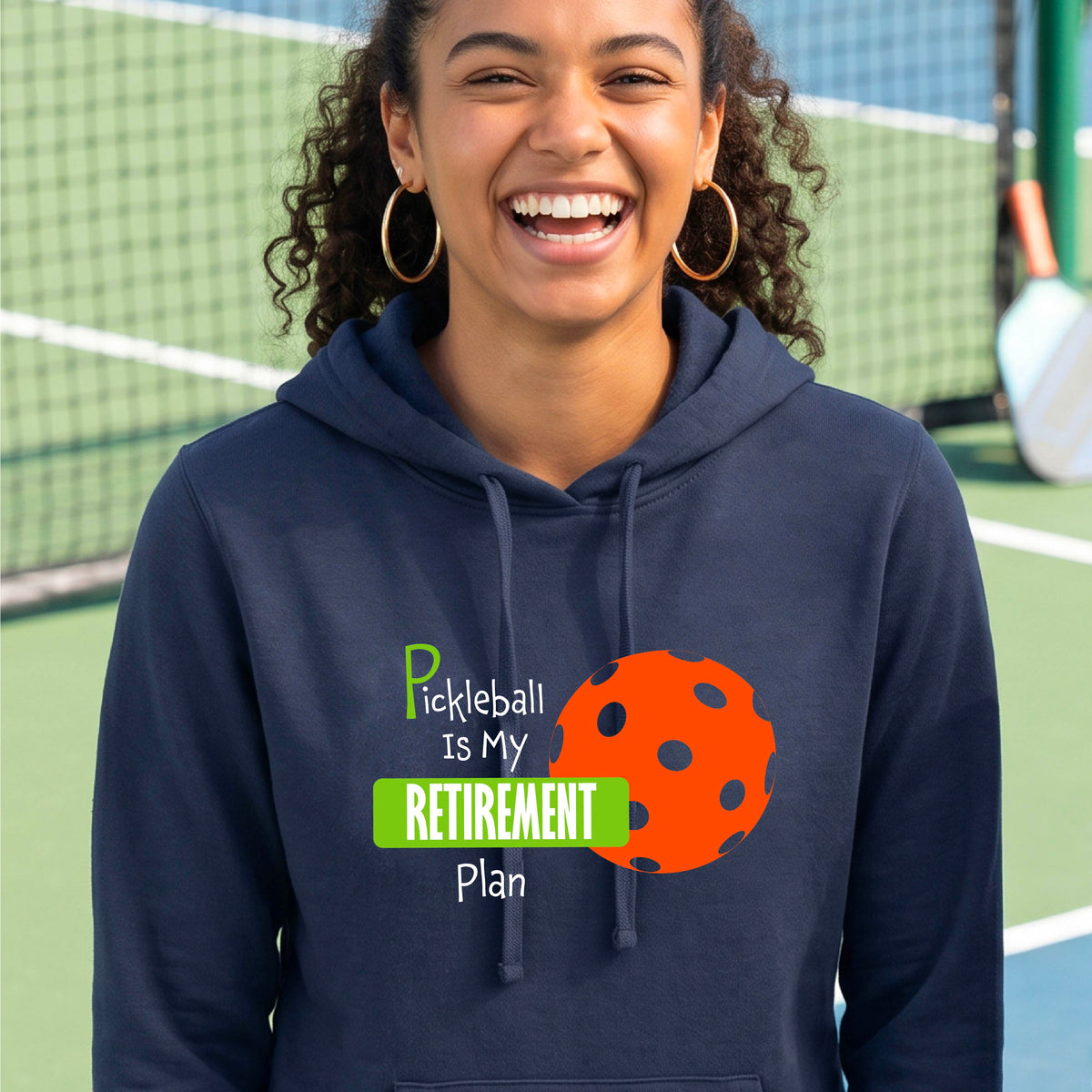 A woman smiling on a pickleball court wearing a navy blue hoodie with a graphic that reads 'Pickleball Is My Retirement Plan' alongside an orange pickleball illustration.