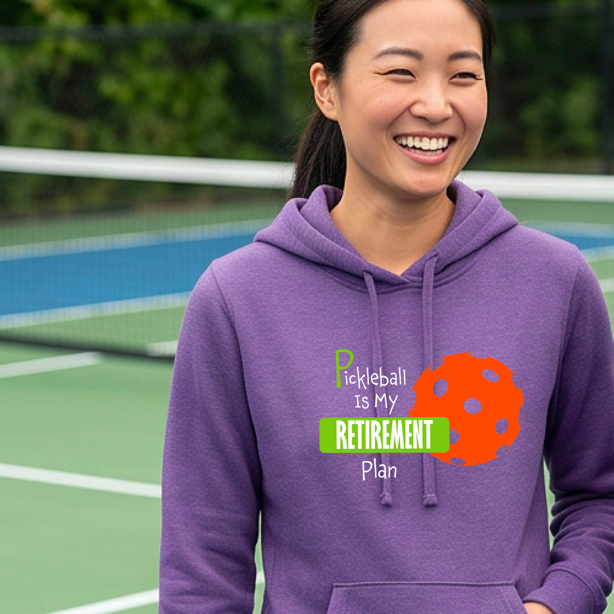 A woman smiling on a pickleball court wearing a purple hoodie with a graphic that reads 'Pickleball Is My Retirement Plan' alongside an orange pickleball illustration.