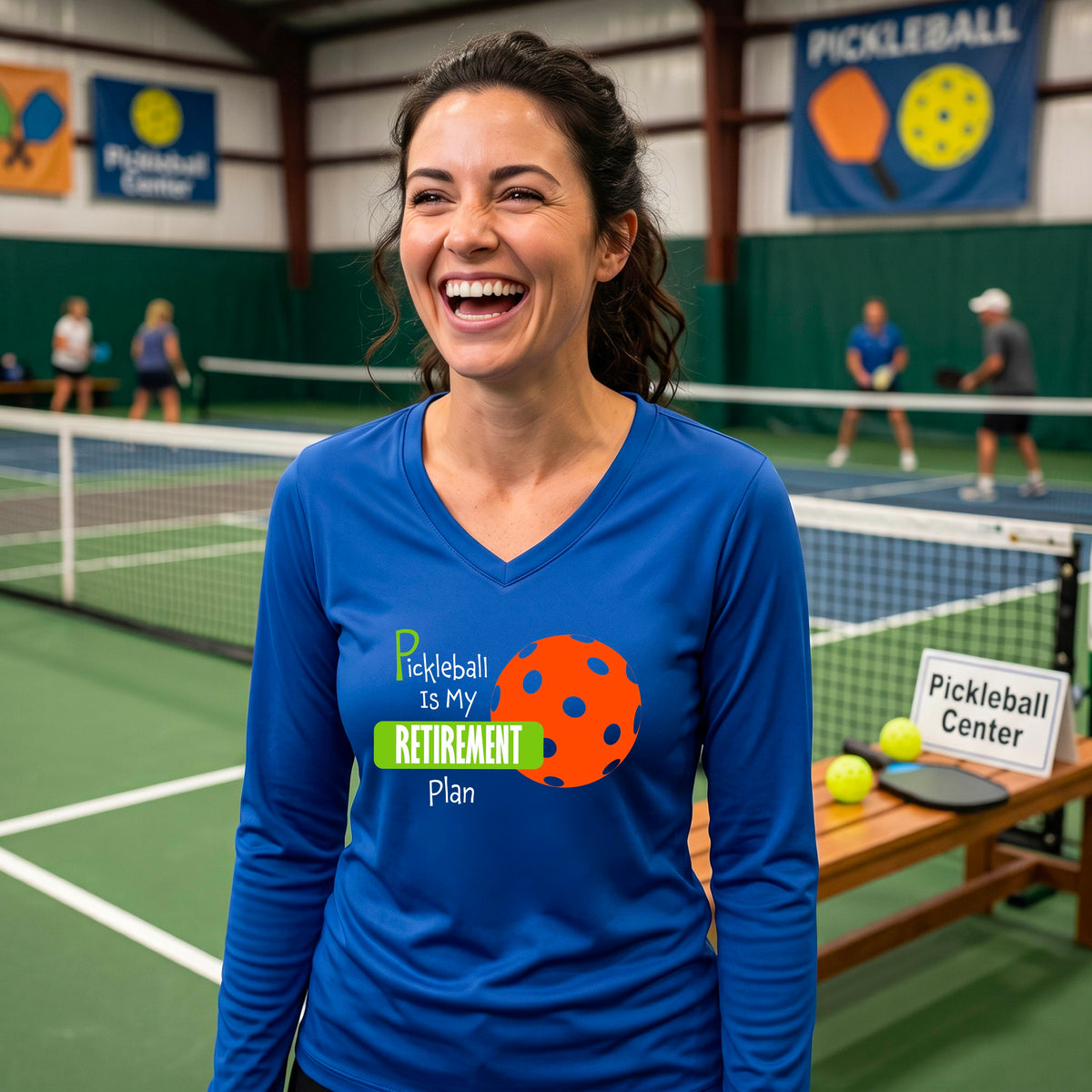 A woman on a pickleball court, wearing a royal blue long-sleeve v-neck athletic t-shirt, with the words “Pickleball is my Retirement Plan” in green and white with an orange pickleball illustration in the background
