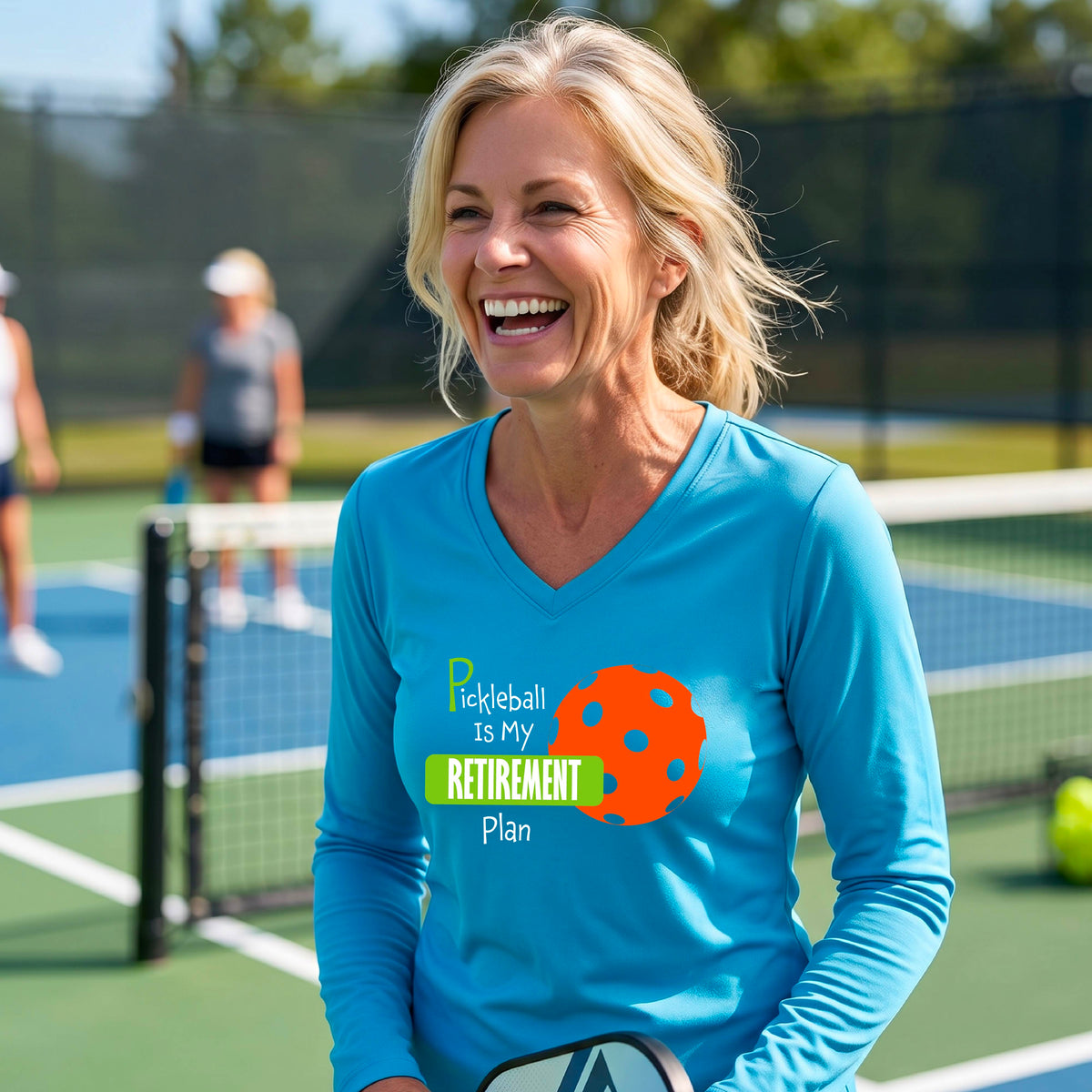 A woman on a pickleball court, wearing a bright blue long-sleeve v-neck athletic t-shirt, with the words “Pickleball is my Retirement Plan” in green and white with an orange pickleball illustration in the background