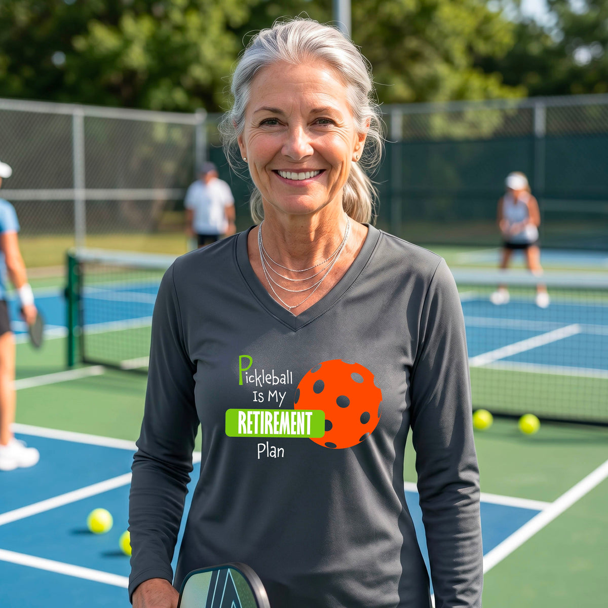 A woman on a pickleball court, wearing a dark gray long-sleeve v-neck athletic t-shirt, with the words “Pickleball is my Retirement Plan” in green and white with an orange pickleball illustration in the background
