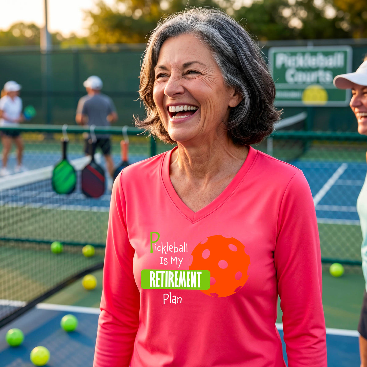 A woman on a pickleball court, wearing a bright coral pink long-sleeve v-neck athletic t-shirt, with the words “Pickleball is my Retirement Plan” in green and white with an orange pickleball illustration in the background