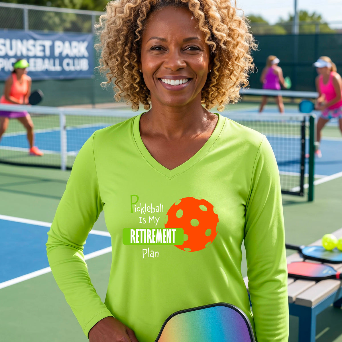 A woman on a pickleball court, wearing a lime green long-sleeve v-neck athletic t-shirt, with the words “Pickleball is my Retirement Plan” in green and white with an orange pickleball illustration in the background