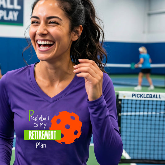 A woman on a pickleball court, wearing a purple long-sleeve v-neck athletic t-shirt, with the words “Pickleball is my Retirement Plan” in green and white with an orange pickleball illustration in the background