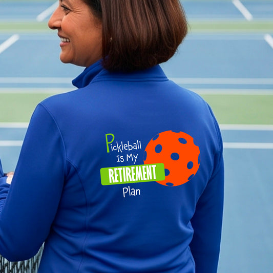 A woman on a pickleball court, showing the back of a royal blue quarter-zip shirt with the words “Pickleball is my Retirement Plan” in green and white with an orange pickleball illustration in the background.