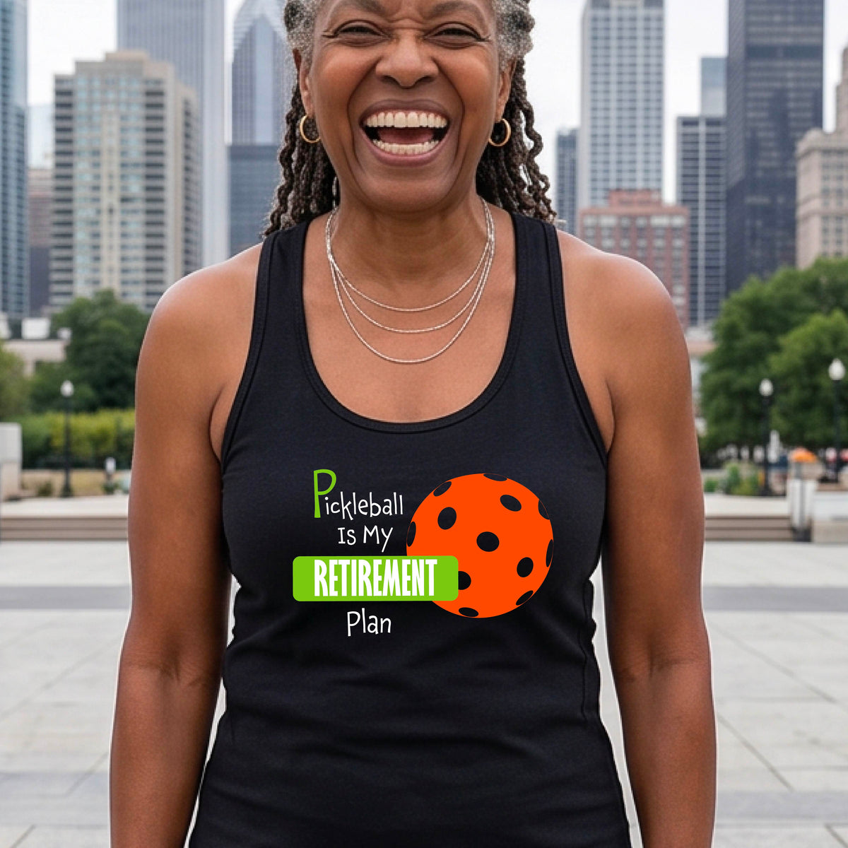 A smiling woman on a pickleball court wearing a black tank top with the text 'Pickleball is My Retirement Plan' and an orange pickleball graphic.