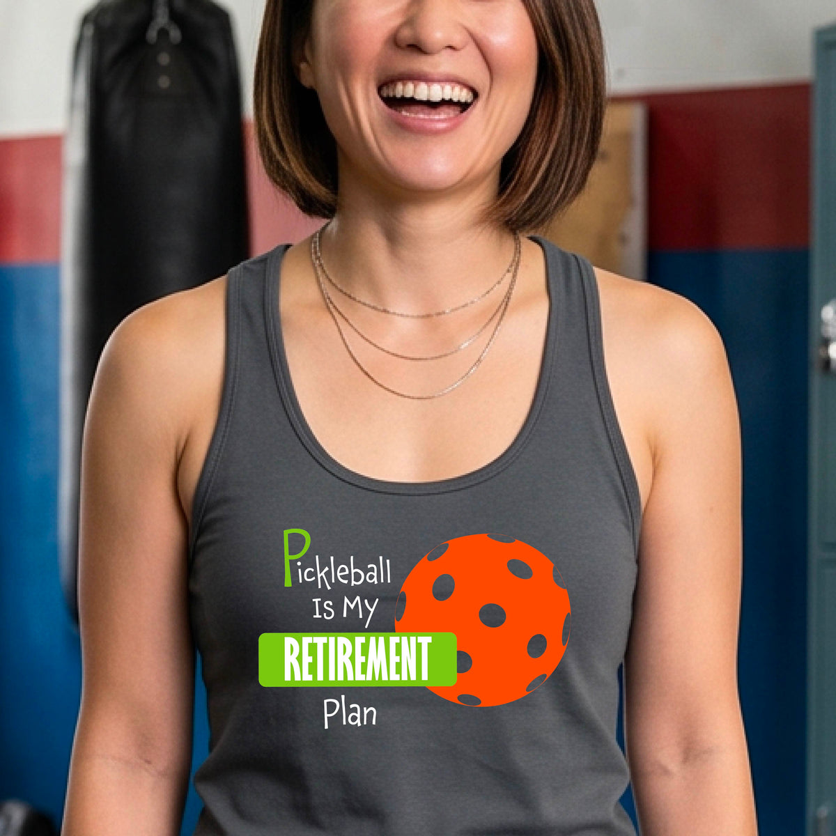 A smiling woman on a pickleball court wearing a dark gray tank top with the text 'Pickleball is My Retirement Plan' and an orange pickleball graphic.
