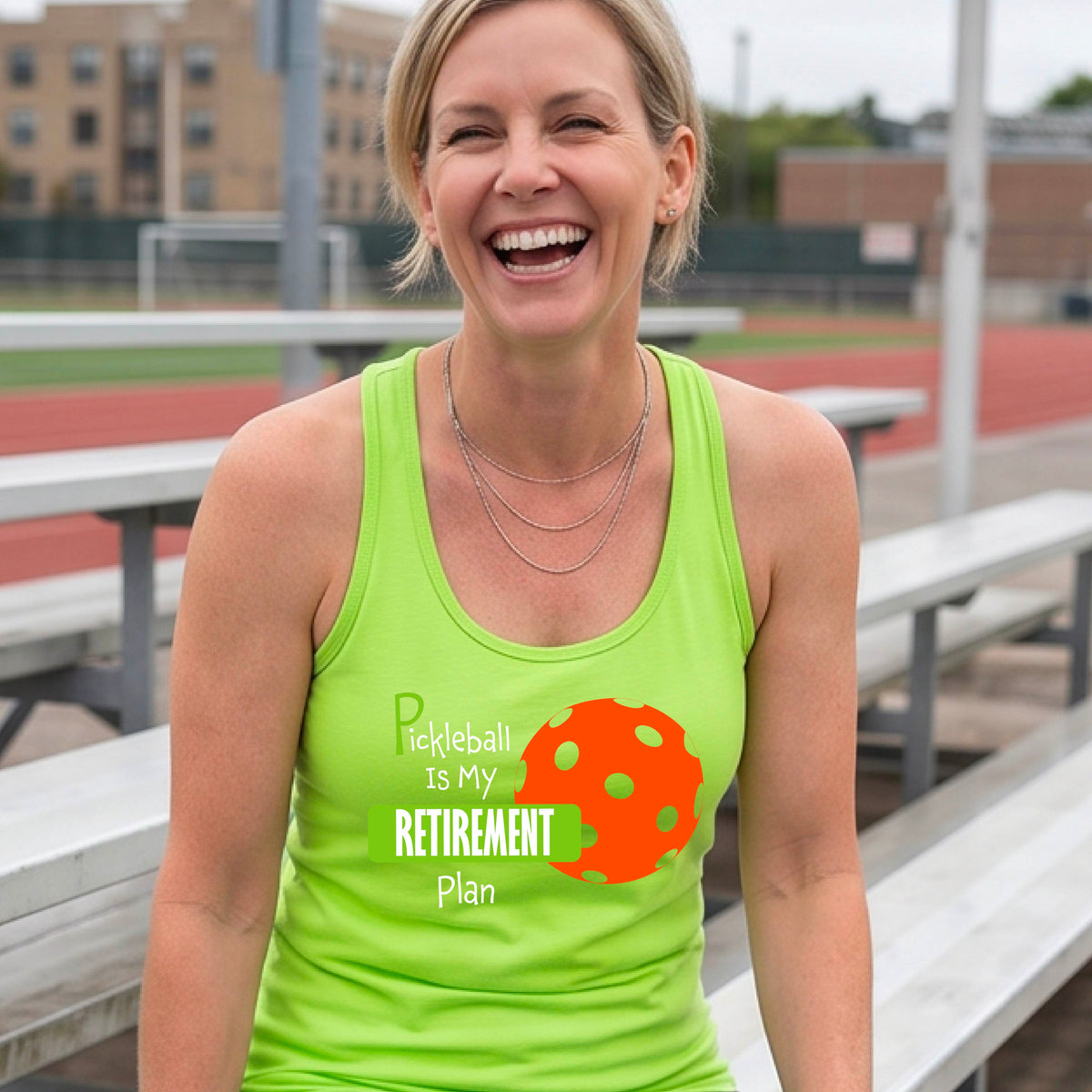 A smiling woman on a pickleball court wearing a lime green tank top with the text 'Pickleball is My Retirement Plan' and an orange pickleball graphic.