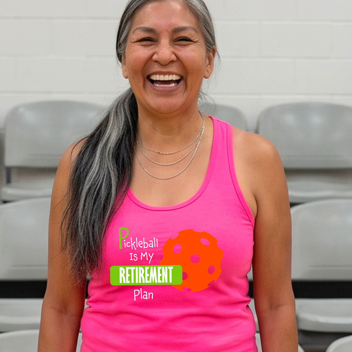 A smiling woman on a pickleball court wearing a pink tank top with the text 'Pickleball is My Retirement Plan' and an orange pickleball graphic.