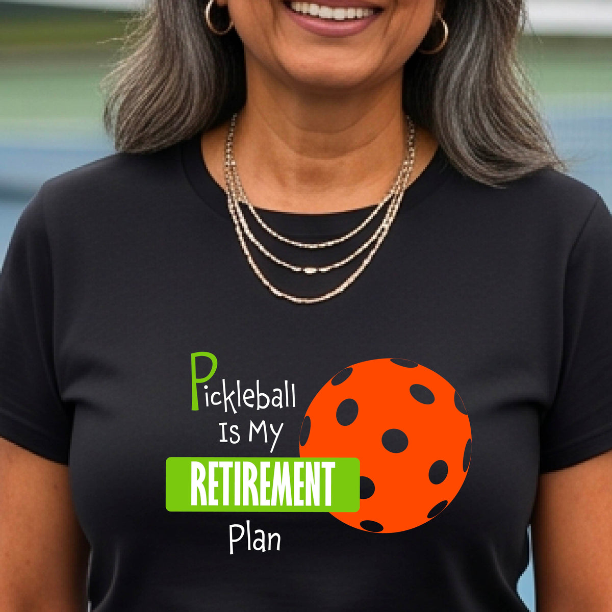 A woman on a pickleball court, wearing a black short-sleeve athletic t-shirt, with the words “Pickleball is my Retirement Plan” in green and white with an orange pickleball illustration in the background