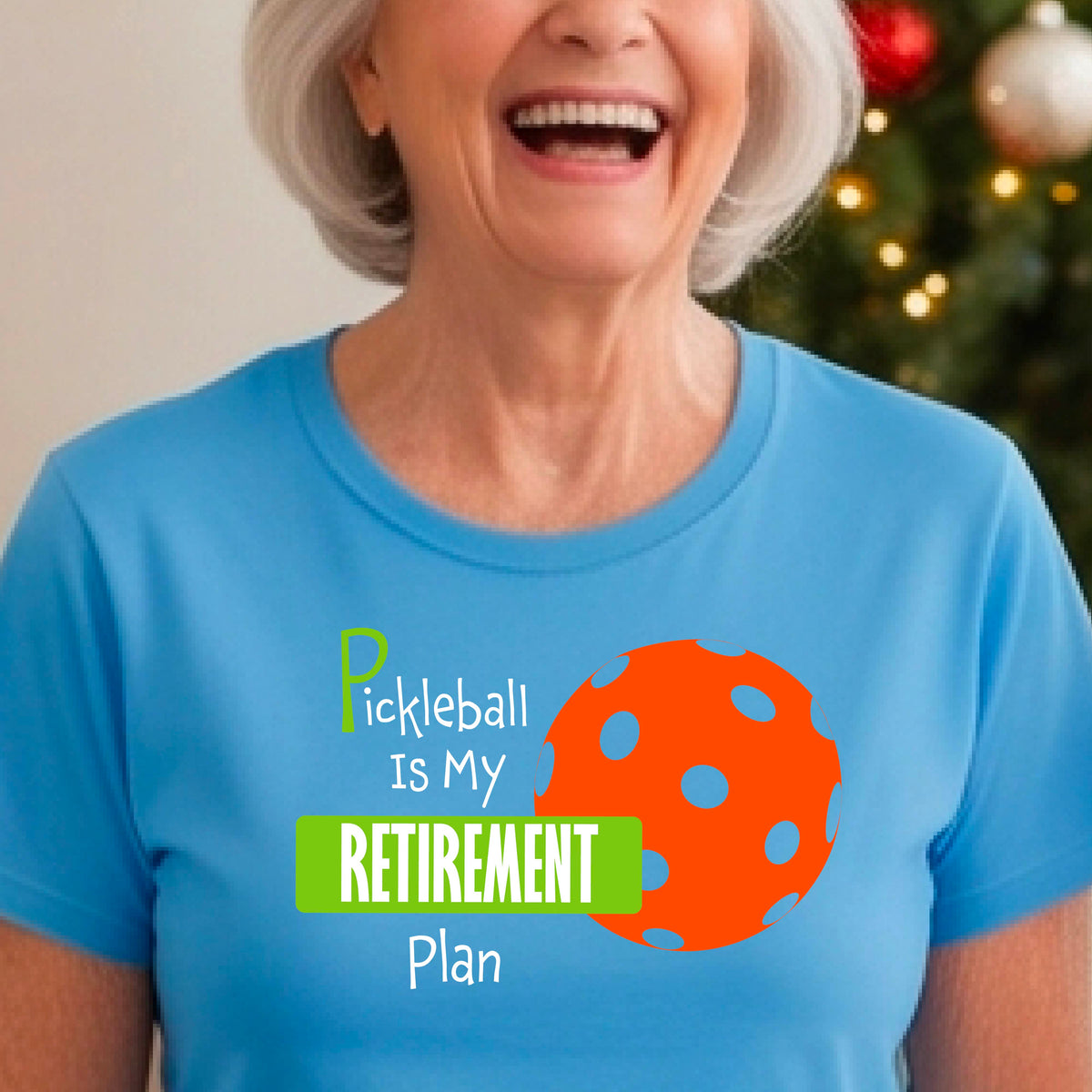 A woman on a pickleball court, wearing a blue short-sleeve athletic t-shirt, with the words “Pickleball is my Retirement Plan” in green and white with an orange pickleball illustration in the background