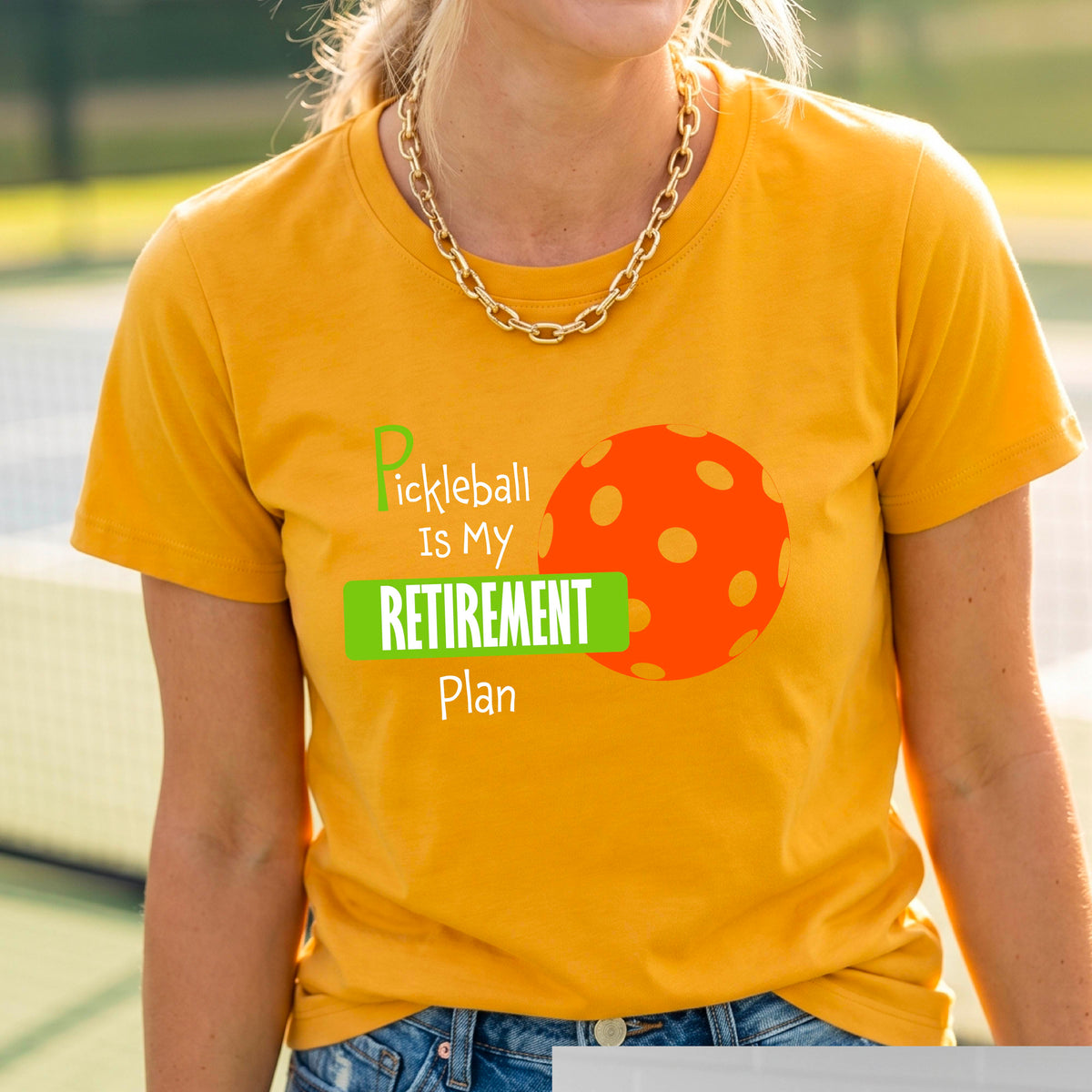 A woman on a pickleball court, wearing a mustard yellow gold  short-sleeve athletic t-shirt, with the words “Pickleball is my Retirement Plan” in green and white with an orange pickleball illustration in the background