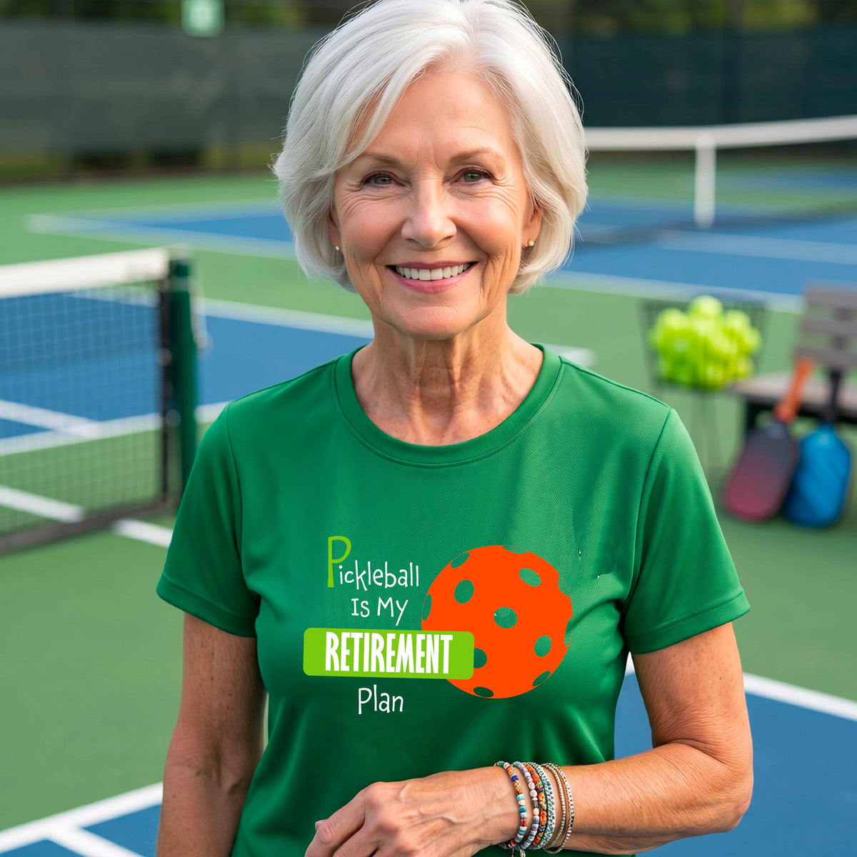 A woman on a pickleball court, wearing a green short-sleeve athletic t-shirt, with the words “Pickleball is my Retirement Plan” in green and white with an orange pickleball illustration in the background