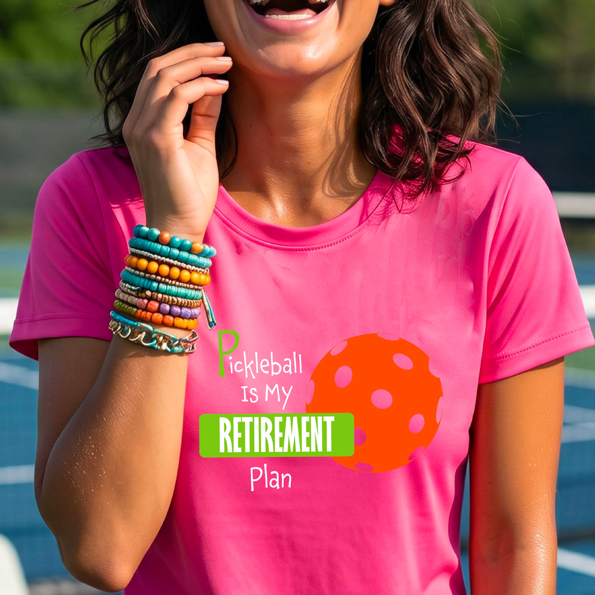 A woman on a pickleball court, wearing a pink short-sleeve athletic t-shirt, with the words “Pickleball is my Retirement Plan” in green and white with an orange pickleball illustration in the background