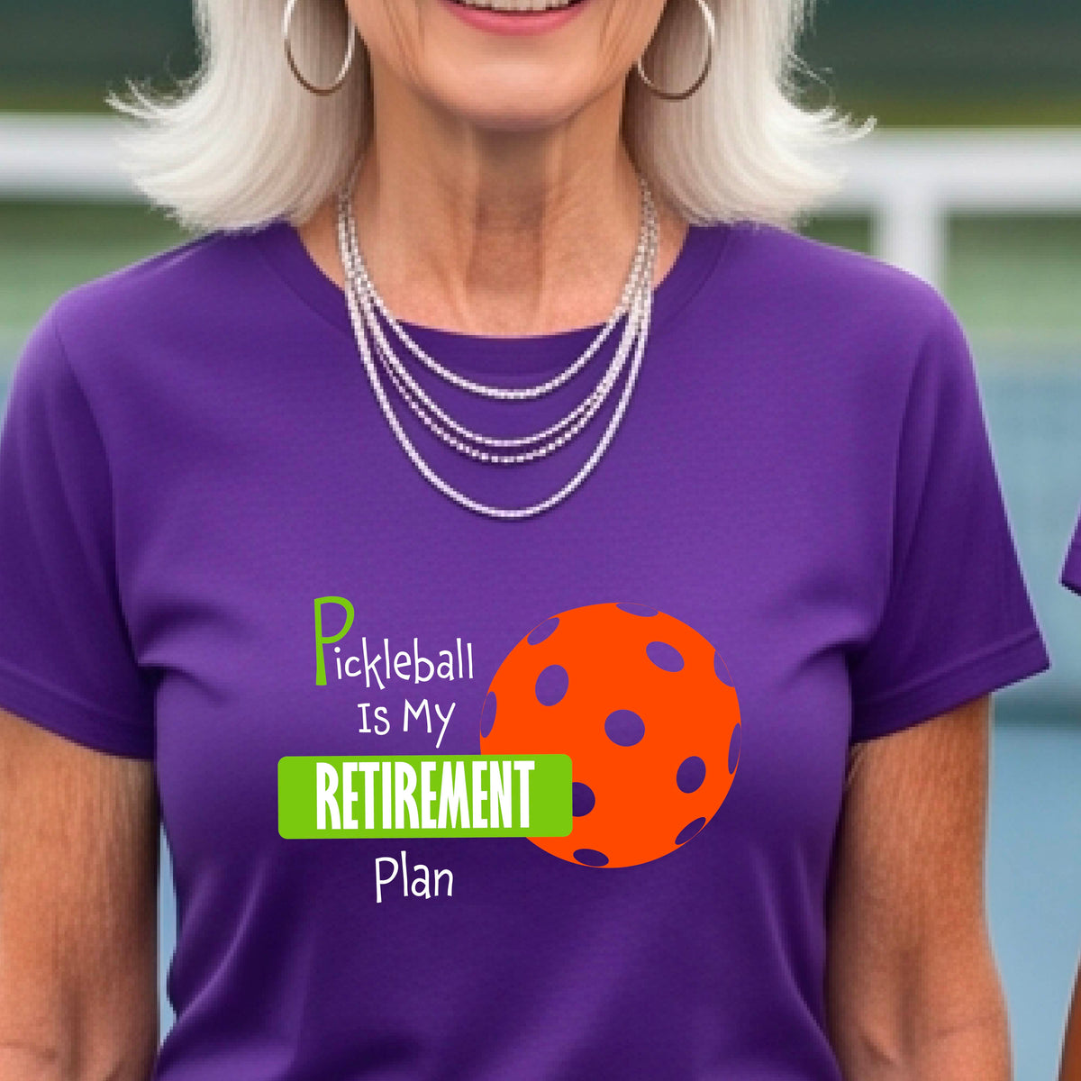 A woman on a pickleball court, wearing a purple short-sleeve athletic t-shirt, with the words “Pickleball is my Retirement Plan” in green and white with an orange pickleball illustration in the background