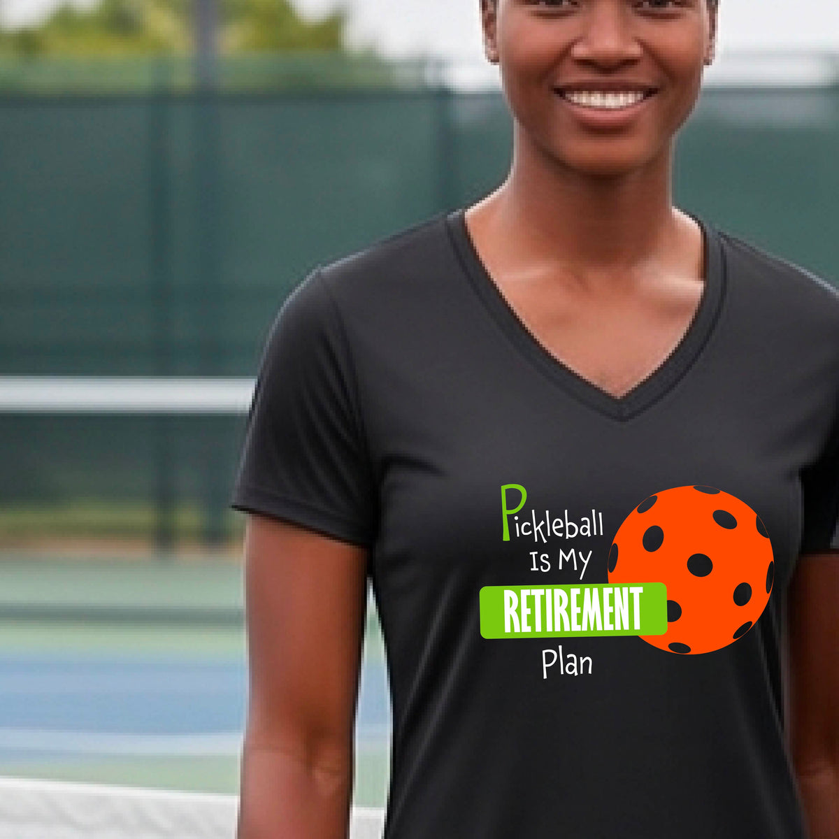 A woman on a pickleball court, wearing a black short-sleeve v-neck athletic t-shirt, with the words “Pickleball is my Retirement Plan” in green and white with an orange pickleball illustration in the background