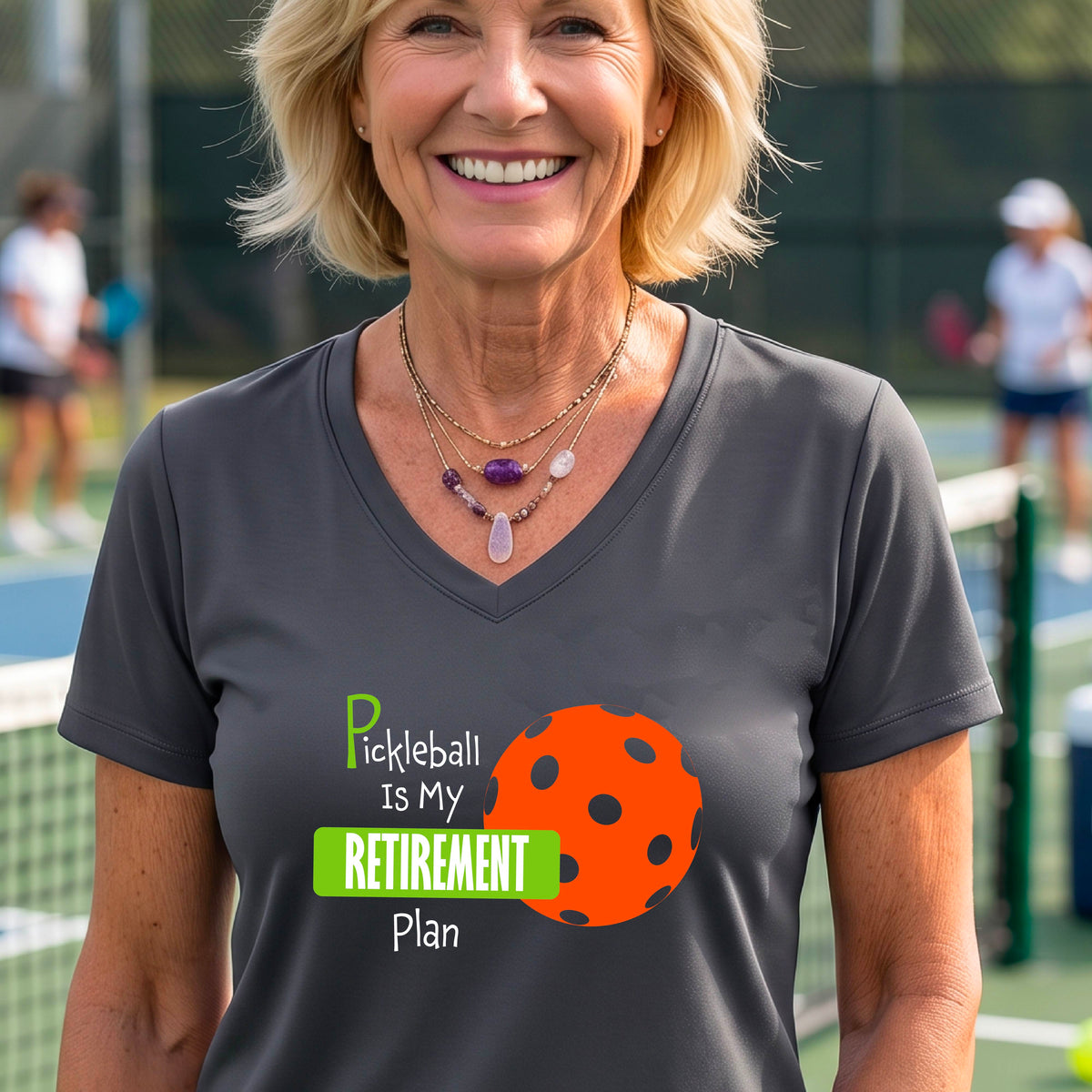A woman on a pickleball court, wearing a gray short-sleeve v-neck athletic t-shirt, with the words “Pickleball is my Retirement Plan” in green and white with an orange pickleball illustration in the background