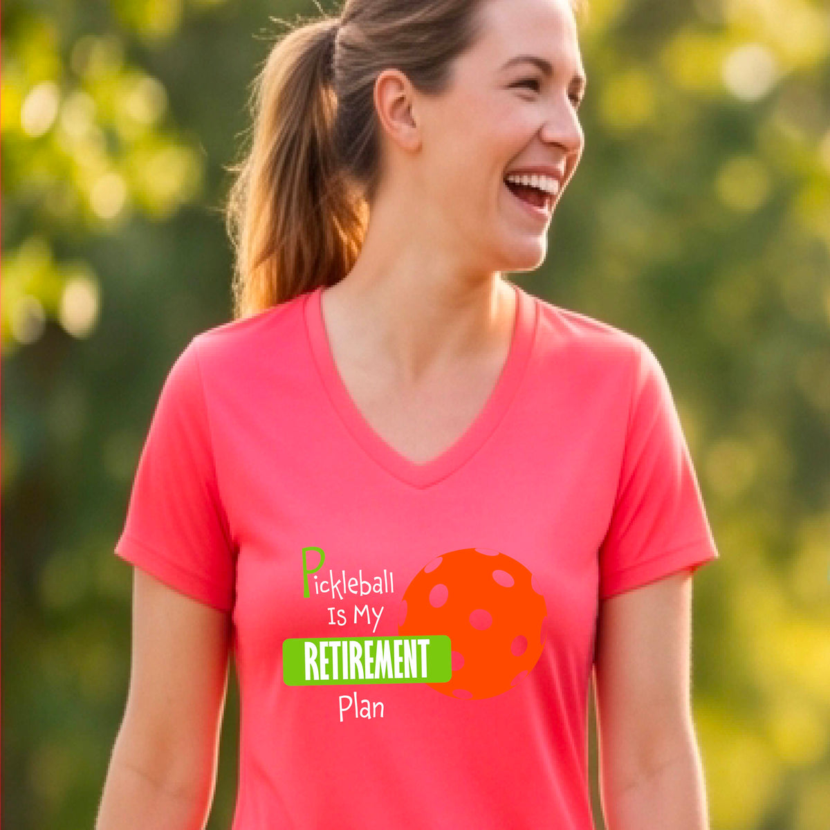 A woman on a pickleball court, wearing a bright coral colored short-sleeve v-neck athletic t-shirt, with the words “Pickleball is my Retirement Plan” in green and white with an orange pickleball illustration in the background