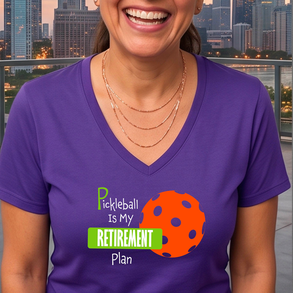 A woman on a pickleball court, wearing a purple short-sleeve v-neck athletic t-shirt, with the words “Pickleball is my Retirement Plan” in green and white with an orange pickleball illustration in the background