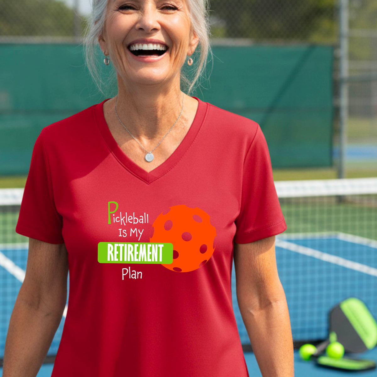 A woman on a pickleball court, wearing a red short-sleeve v-neck athletic t-shirt, with the words “Pickleball is my Retirement Plan” in green and white with an orange pickleball illustration in the background