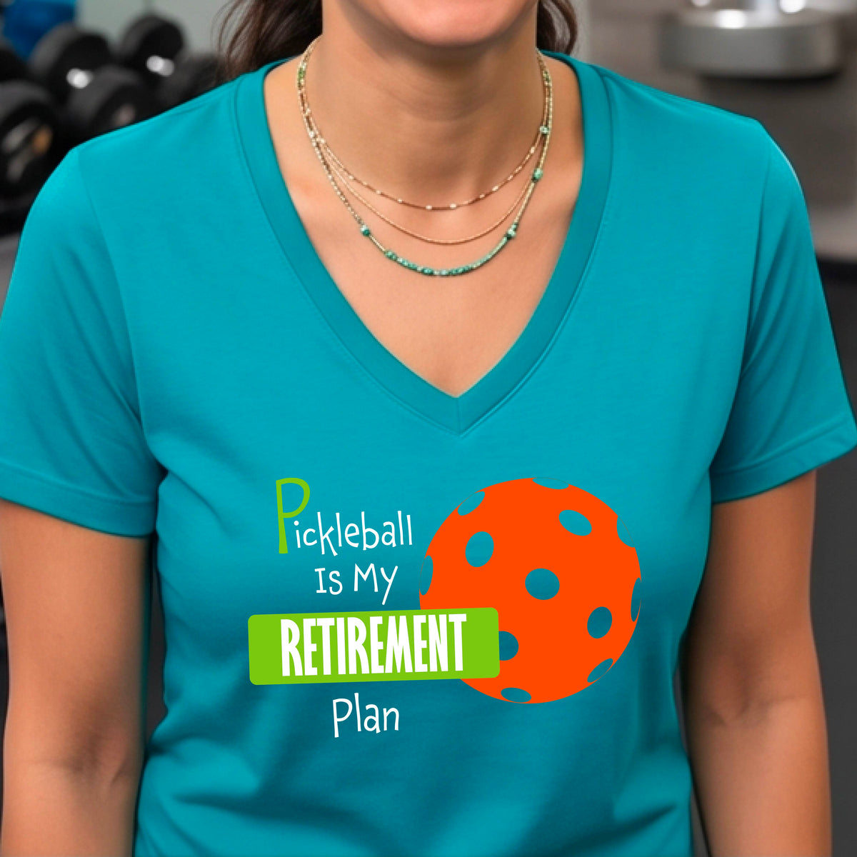A woman on a pickleball court, wearing a teal short-sleeve v-neck athletic t-shirt, with the words “Pickleball is my Retirement Plan” in green and white with an orange pickleball illustration in the background