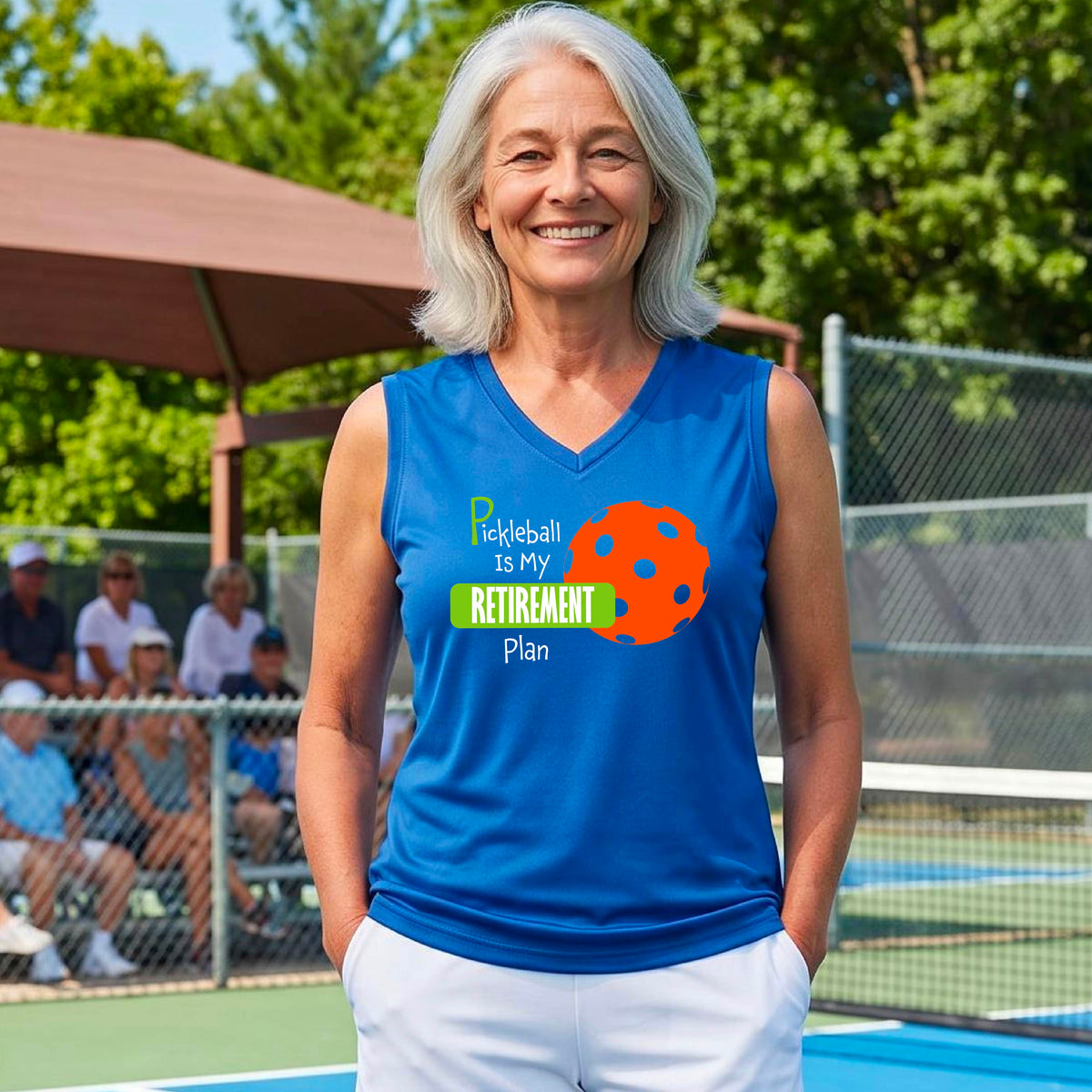 A woman on a pickleball court, wearing a royal blue sleeveless v-neck athletic t-shirt, with the words “Pickleball is my Retirement Plan” in green and white with an orange pickleball illustration in the background