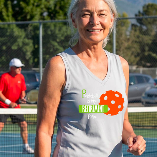 A woman on a pickleball court, wearing a silver gray sleeveless v-neck athletic t-shirt, with the words “Pickleball is my Retirement Plan” in green and white with an orange pickleball illustration in the background