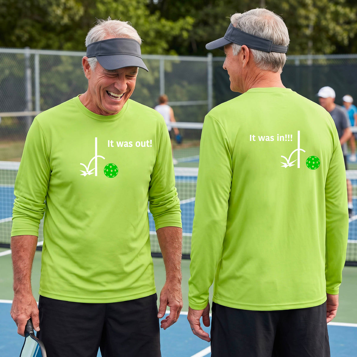 Two men showing a lime green long-sleeve t-shirt with a white design of ‘IT WAS OUT!’ and a colorful pickleball on the front, and the back design with a white ‘IT WAS IN!!!’ and a colorful pickleball, standing on a pickleball court.