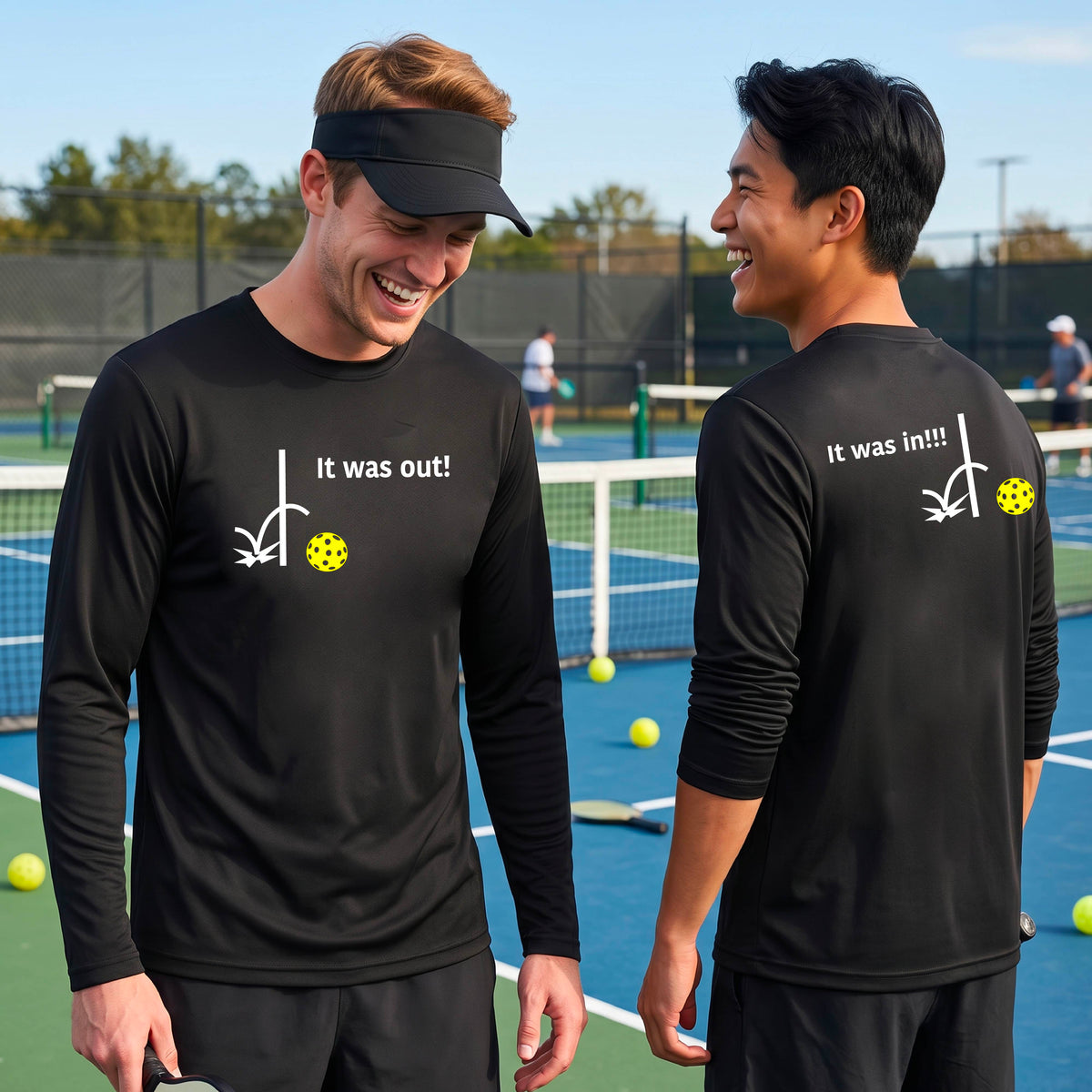 Two men showing a black long-sleeve t-shirt with a white design of ‘IT WAS OUT!’ and a colorful pickleball on the front, and the back design with a white ‘IT WAS IN!!!’ and a colorful pickleball, standing on a pickleball court.