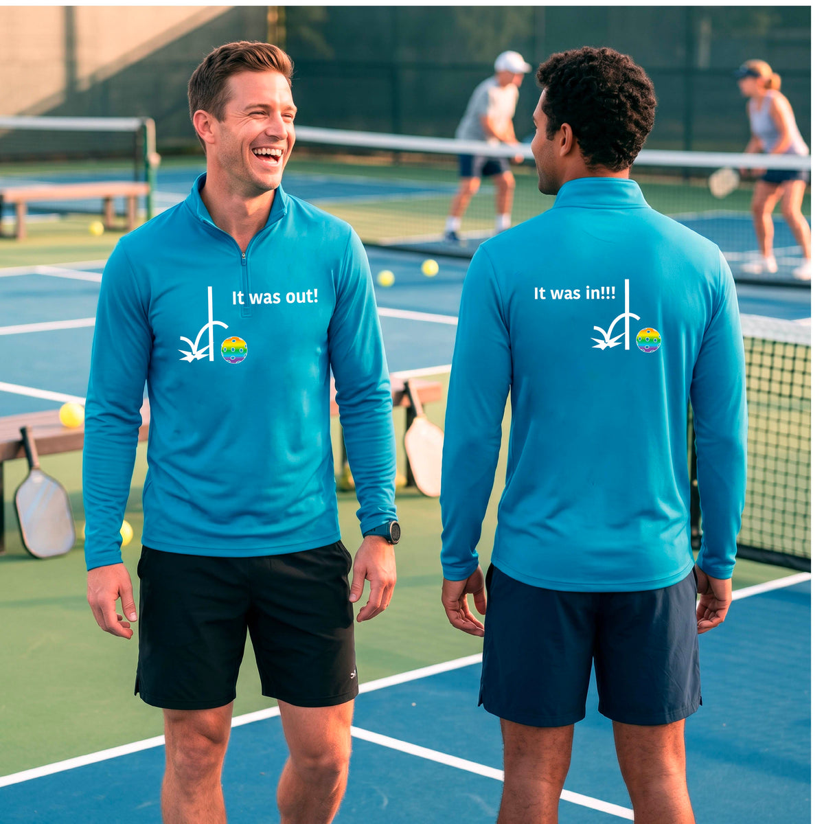 Two men wearing a bright blue quarter-zip shirt with a white design of ‘IT WAS OUT!’ and a colorful pickleball on the front, and the back design with a white ‘IT WAS IN!!!’ and a colorful pickleball, standing on a pickleball court.