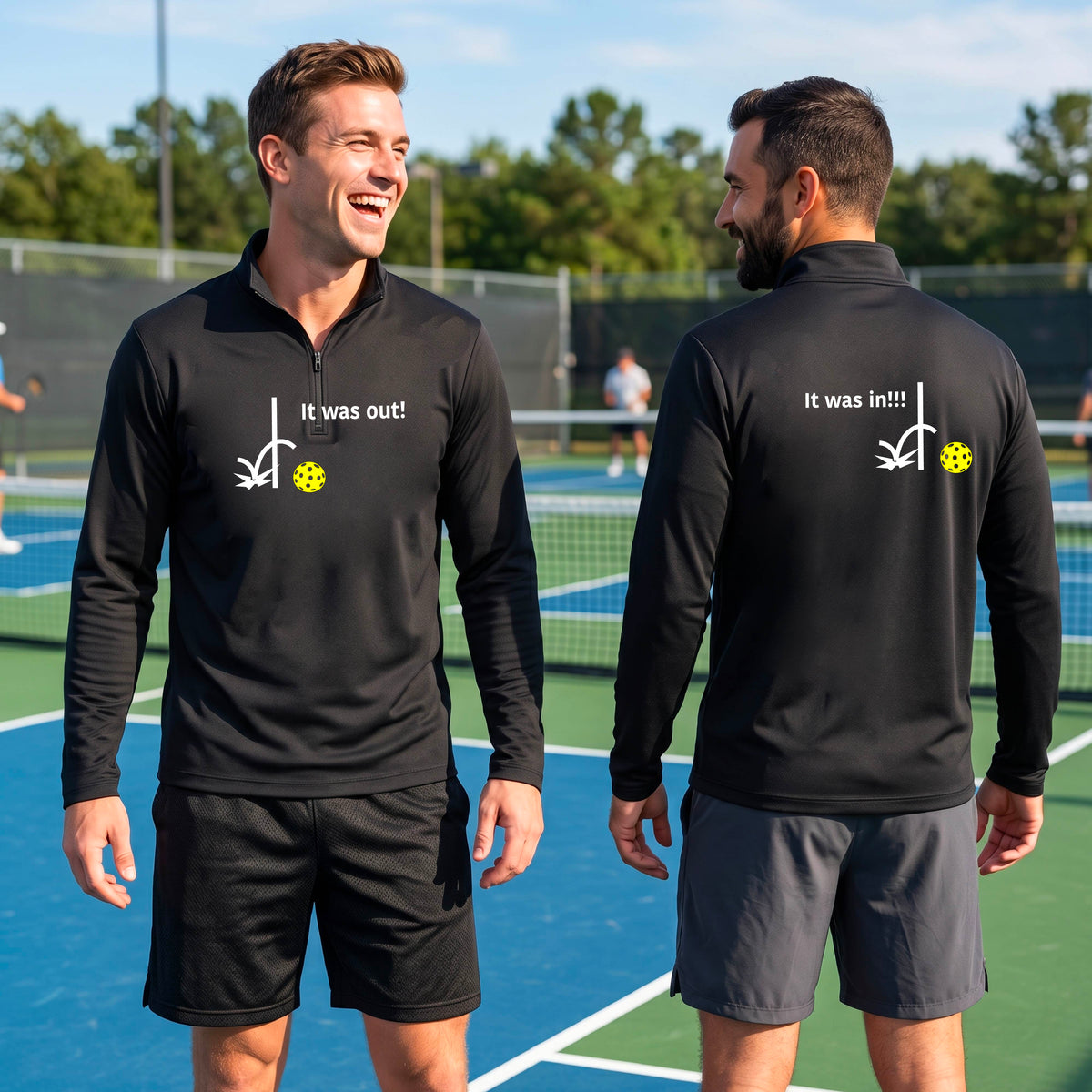 Two men wearing a black quarter-zip shirt with a white design of ‘IT WAS OUT!’ and a colorful pickleball on the front, and the back design with a white ‘IT WAS IN!!!’ and a colorful pickleball, standing on a pickleball court.