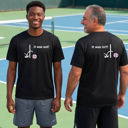 Two men showing a black short-sleeve t-shirt with a white design of ‘IT WAS OUT!’ and a colorful pickleball on the front, and the back design with a white ‘IT WAS IN!!!’ and a colorful pickleball, standing on a pickleball court. 