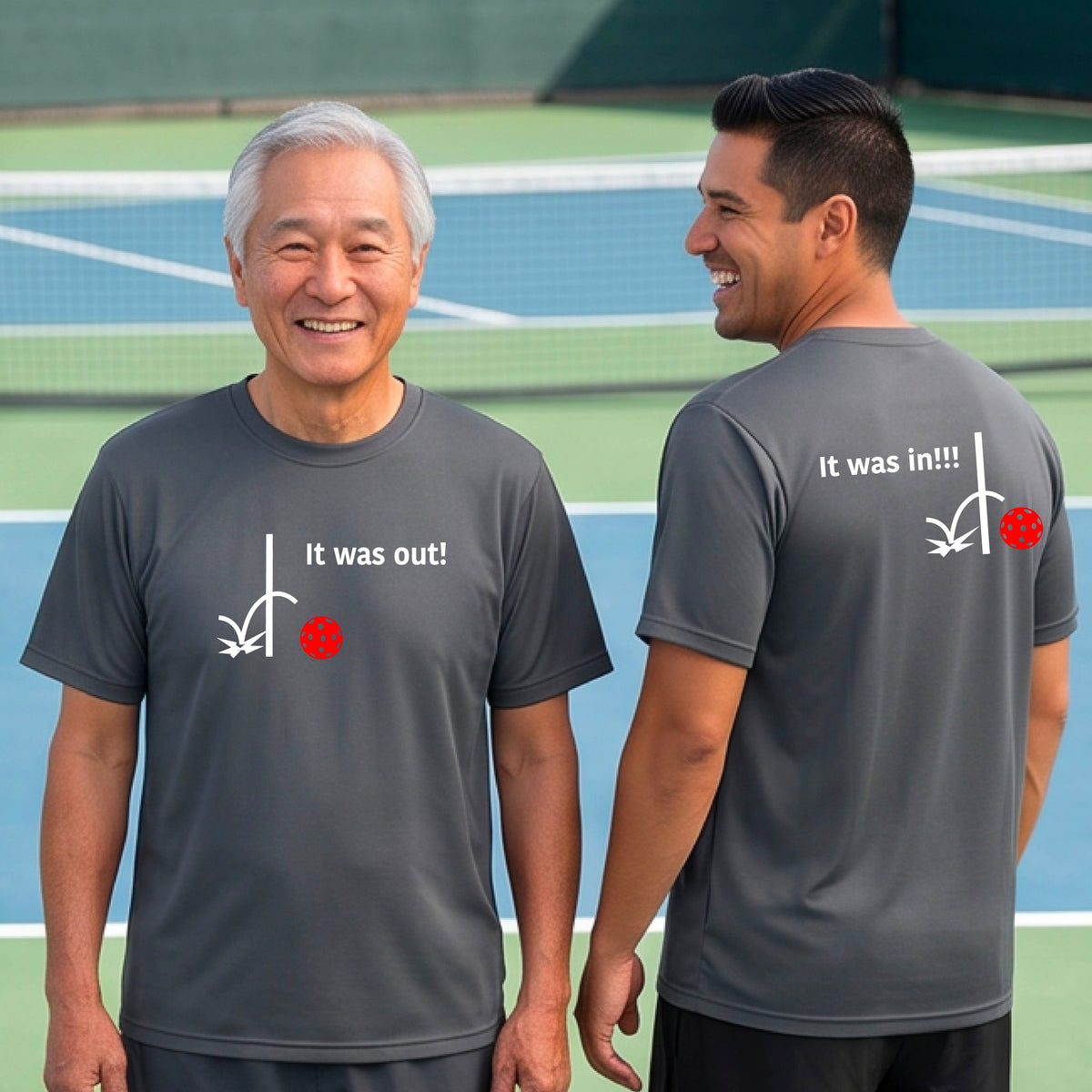 Two men showing a gray short-sleeve t-shirt with a white design of ‘IT WAS OUT!’ and a colorful pickleball on the front, and the back design with a white ‘IT WAS IN!!!’ and a colorful pickleball, standing on a pickleball court. 