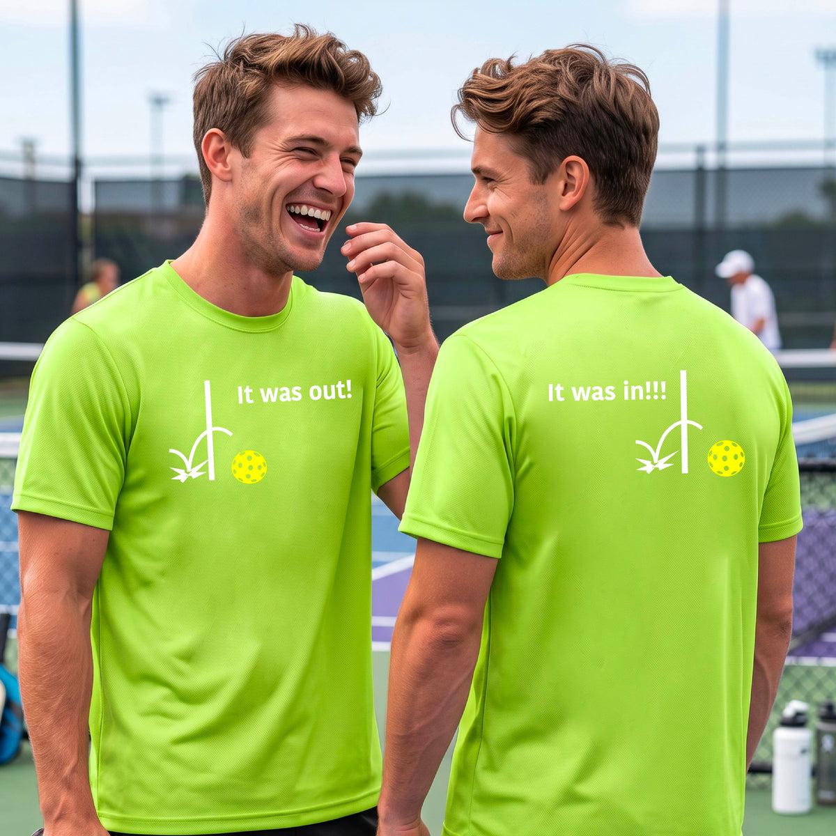 Two men showing a lime green short-sleeve t-shirt with a white design of ‘IT WAS OUT!’ and a colorful pickleball on the front, and the back design with a white ‘IT WAS IN!!!’ and a colorful pickleball, standing on a pickleball court. 
