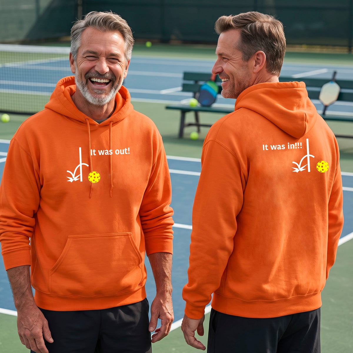 Two men showing an orange hoodie pullover with a white design of ‘IT WAS OUT!’ and a colorful pickleball on the front, and the back design with a white ‘IT WAS IN!!!’ and a colorful pickleball, standing on a pickleball court.
