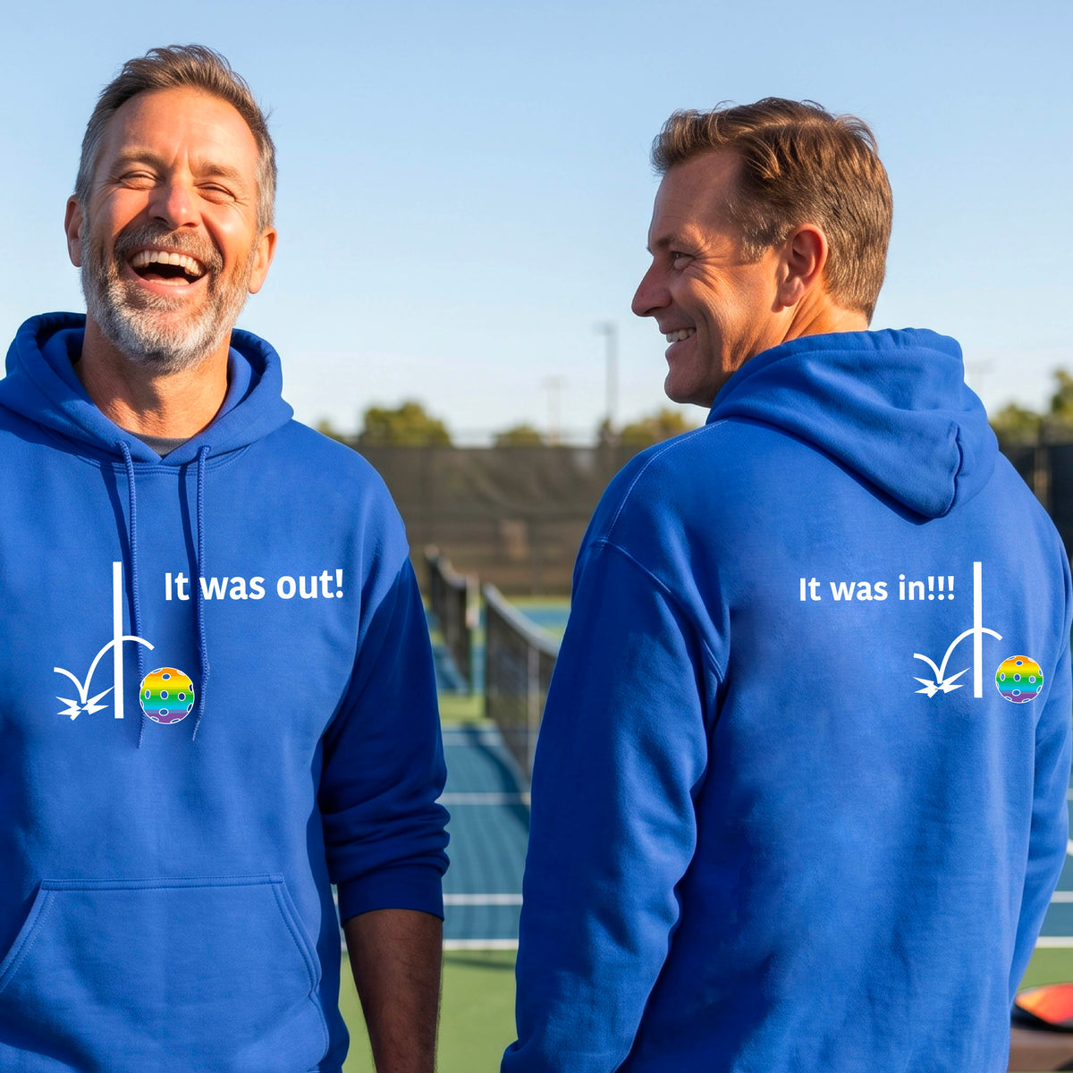 Two men showing a royal blue hoodie pullover with a white design of ‘IT WAS OUT!’ and a colorful pickleball on the front, and the back design with a white ‘IT WAS IN!!!’ and a colorful pickleball, standing on a pickleball court.
