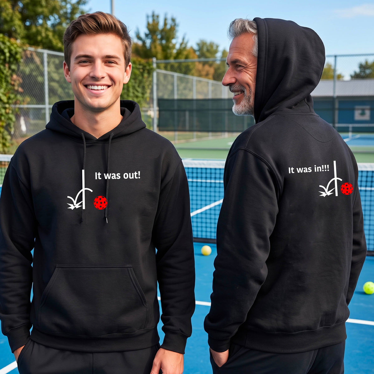 Two men showing a black hoodie pullover with a white design of ‘IT WAS OUT!’ and a colorful pickleball on the front, and the back design with a white ‘IT WAS IN!!!’ and a colorful pickleball, standing on a pickleball court.
