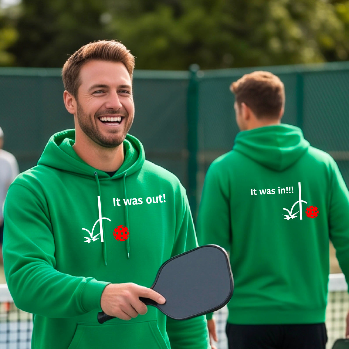 Two men showing a green hoodie pullover with a white design of ‘IT WAS OUT!’ and a colorful pickleball on the front, and the back design with a white ‘IT WAS IN!!!’ and a colorful pickleball, standing on a pickleball court.
