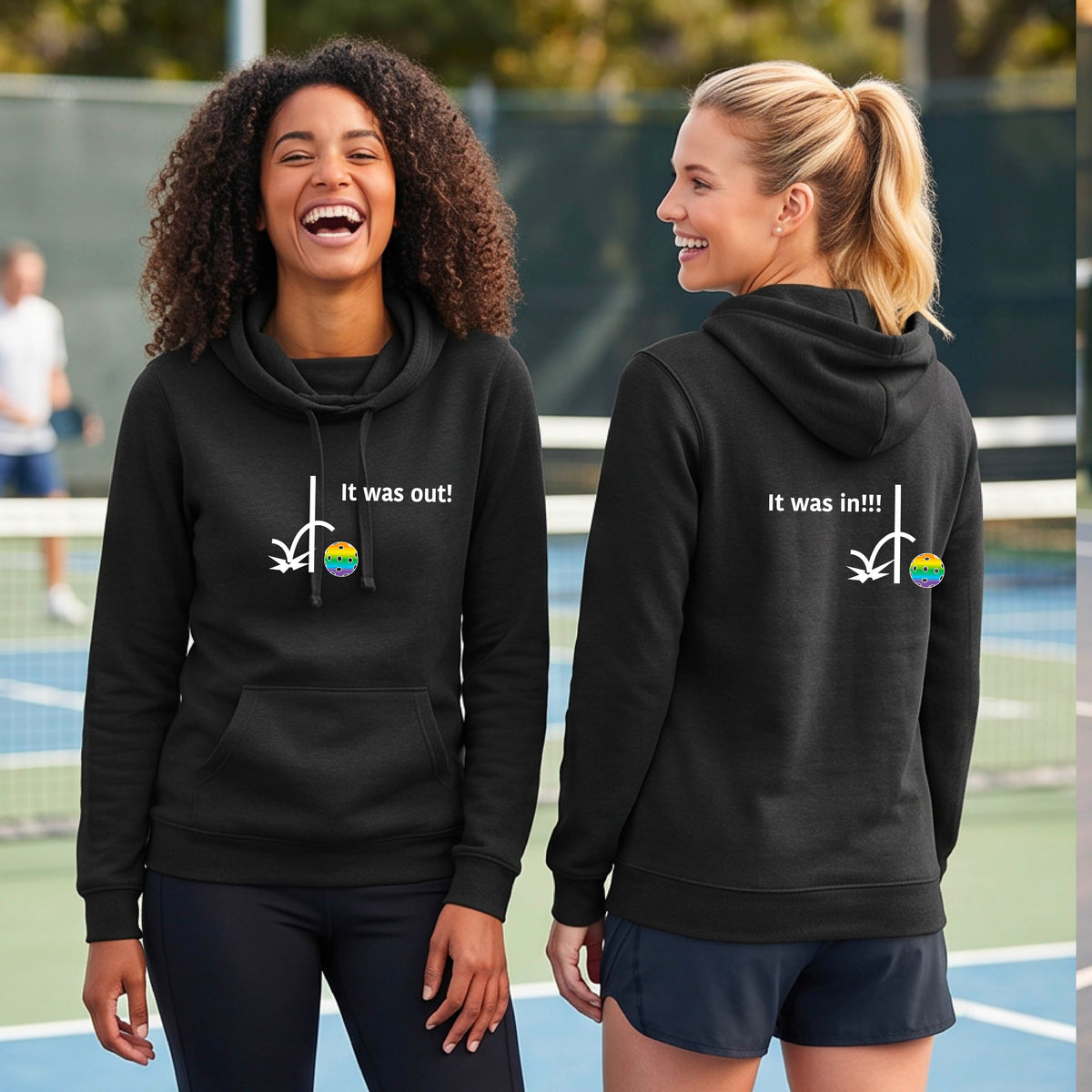 Two women wearing a black cowl-neck fitted hoodie pullover t with a white design of ‘IT WAS OUT!’ and a colorful pickleball on the front, and the back design with a white ‘IT WAS IN!!!’ and a colorful pickleball, standing on a pickleball court.