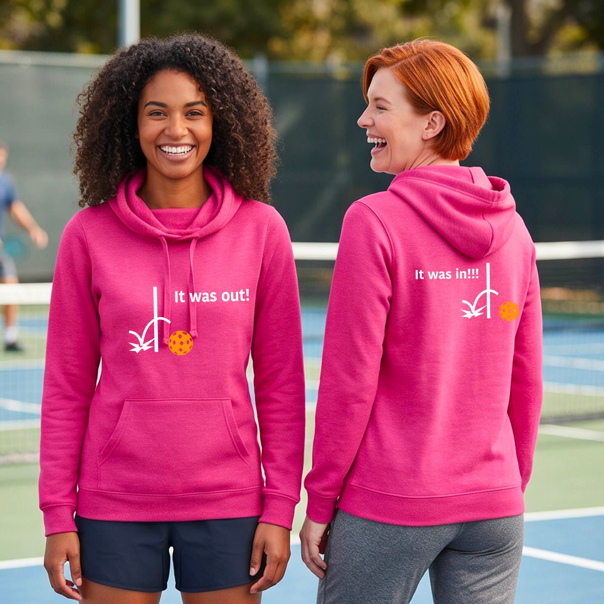 Two women wearing a pink cowl-neck fitted hoodie pullover t with a white design of ‘IT WAS OUT!’ and a colorful pickleball on the front, and the back design with a white ‘IT WAS IN!!!’ and a colorful pickleball, standing on a pickleball court.