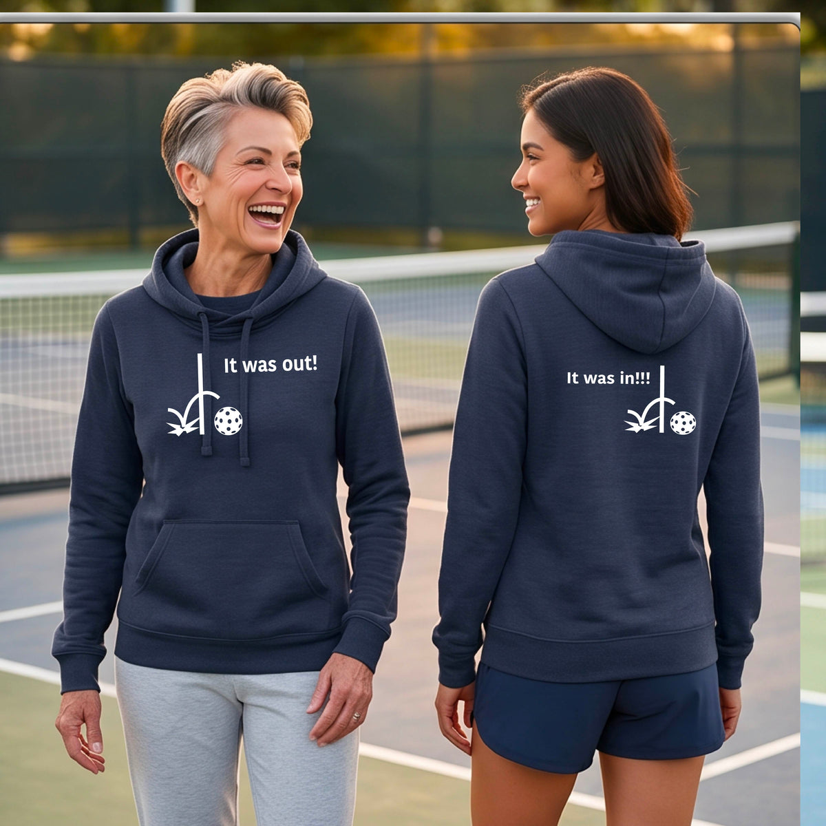 Two women wearing a navy blue Cowl-neck fitted hoodie pullover t with a white design of ‘IT WAS OUT!’ and a colorful pickleball on the front, and the back design with a white ‘IT WAS IN!!!’ and a colorful pickleball, standing on a pickleball court.
