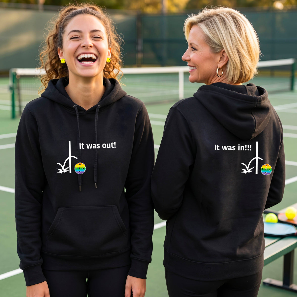 Two women wearing a black fitted hoodie pullover with a white design of ‘IT WAS OUT!’ and a colorful pickleball on the front, and the back design with a white ‘IT WAS IN!!!’ and a colorful pickleball, standing on a pickleball court.