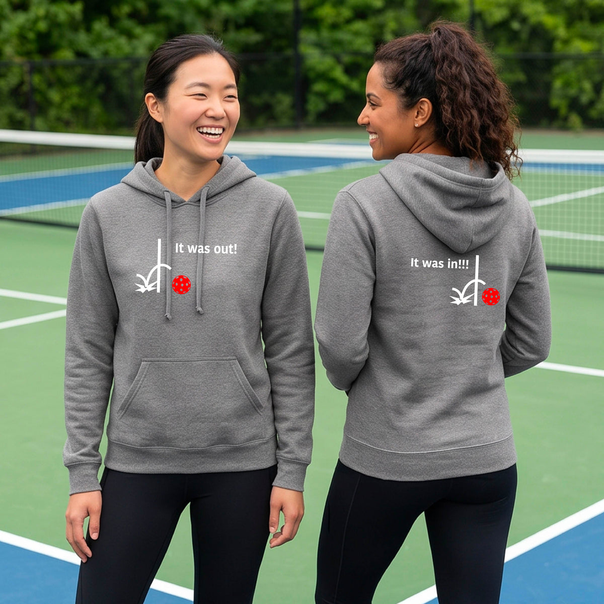 Two women wearing a light gray fitted hoodie pullover with a white design of ‘IT WAS OUT!’ and a colorful pickleball on the front, and the back design with a white ‘IT WAS IN!!!’ and a colorful pickleball, standing on a pickleball court.
