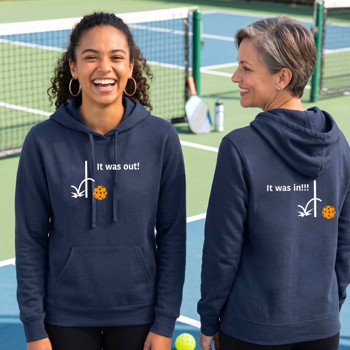 Two women wearing a navy blue fitted hoodie pullover with a white design of ‘IT WAS OUT!’ and a colorful pickleball on the front, and the back design with a white ‘IT WAS IN!!!’ and a colorful pickleball, standing on a pickleball court.