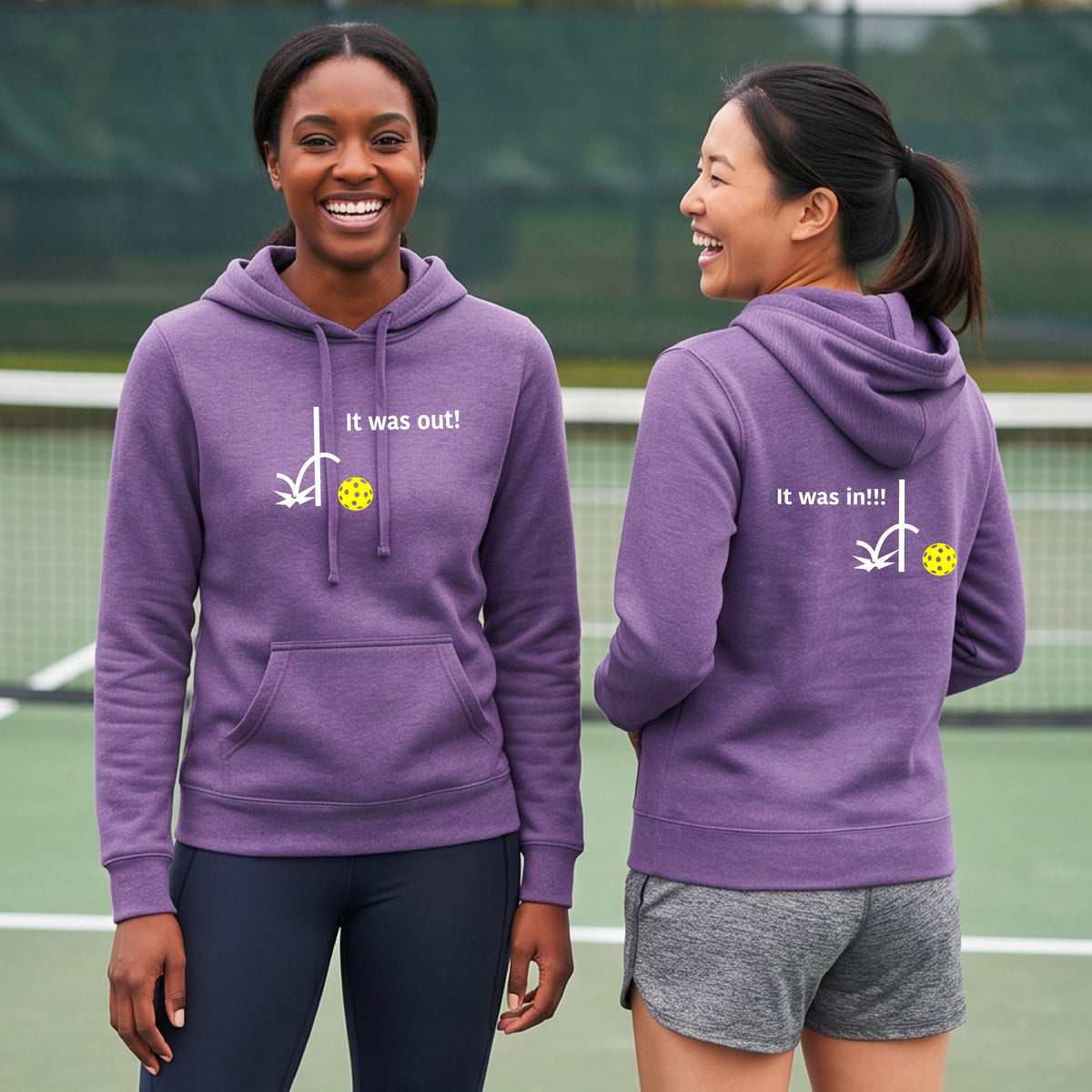 Two women wearing a purple fitted hoodie pullover with a white design of ‘IT WAS OUT!’ and a colorful pickleball on the front, and the back design with a white ‘IT WAS IN!!!’ and a colorful pickleball, standing on a pickleball court.