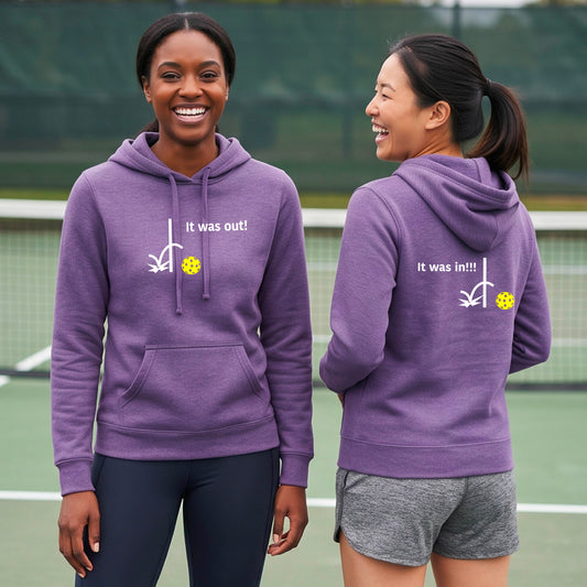 Two women wearing a purple fitted hoodie pullover with a white design of ‘IT WAS OUT!’ and a colorful pickleball on the front, and the back design with a white ‘IT WAS IN!!!’ and a colorful pickleball, standing on a pickleball court.