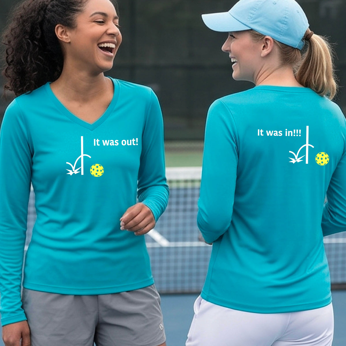 Two women wearing a bright blue long-sleeve t-shirt with a white design of ‘IT WAS OUT!’ and a colorful pickleball on the front, and the back design with a white ‘IT WAS IN!!!’ and a colorful pickleball, standing on a pickleball court.