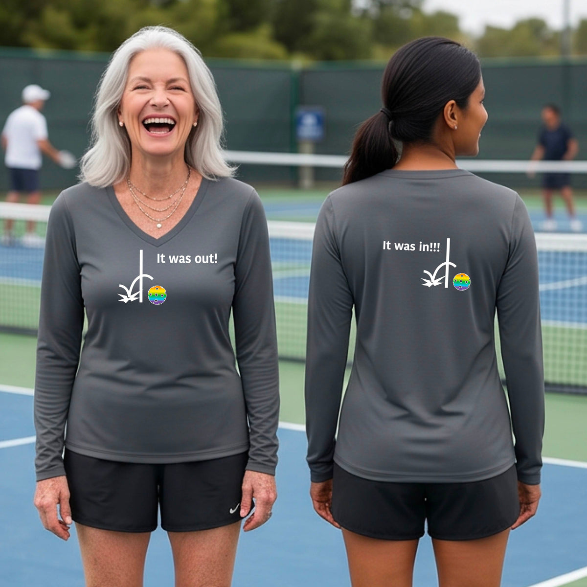 Two women wearing a dark gray long-sleeve t-shirt with a white design of ‘IT WAS OUT!’ and a colorful pickleball on the front, and the back design with a white ‘IT WAS IN!!!’ and a colorful pickleball, standing on a pickleball court.