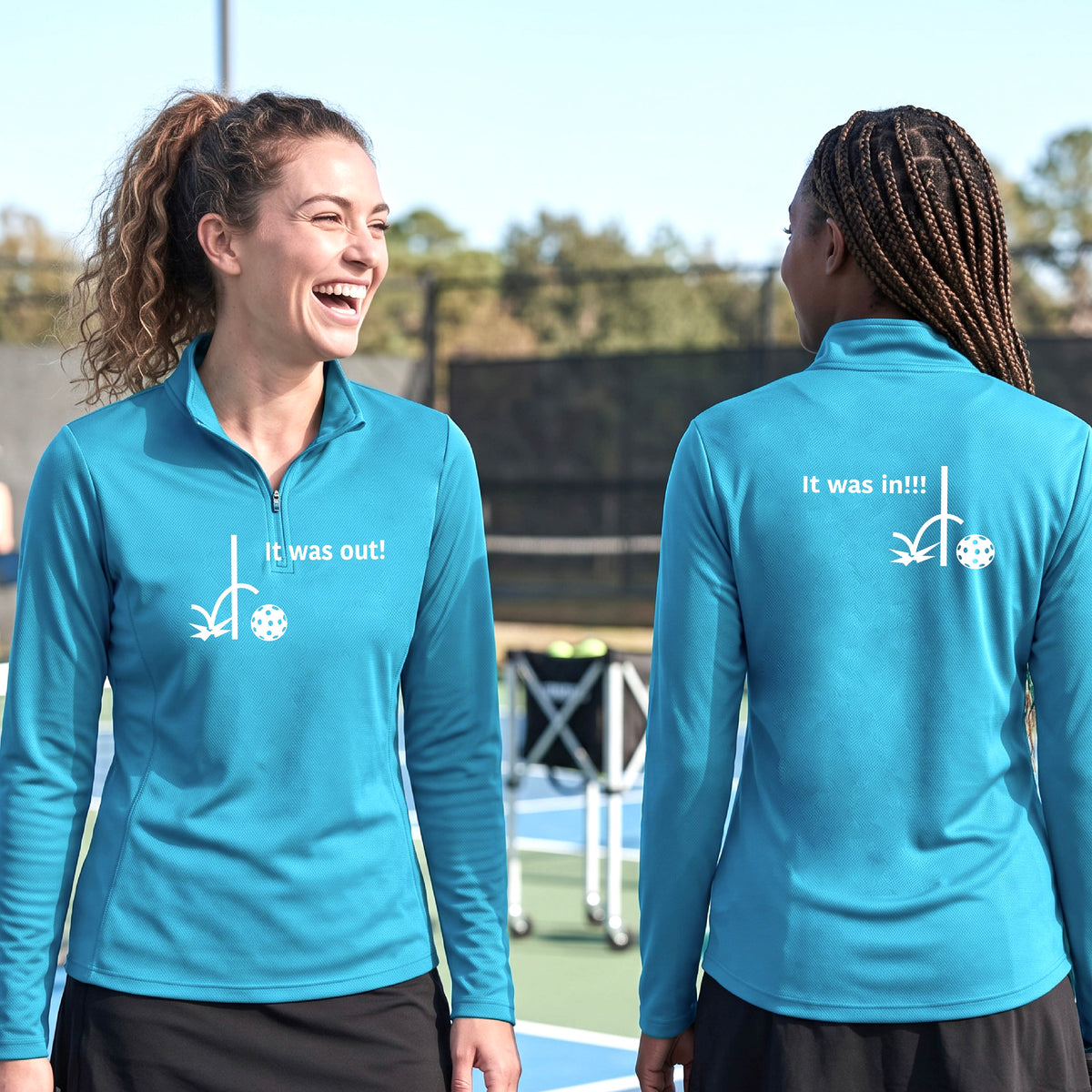 Two women wearing a bright blue quarter-zip shirt with a white design of ‘IT WAS OUT!’ and a colorful pickleball on the front, and the back design with a white ‘IT WAS IN!!!’ and a colorful pickleball, standing on a pickleball court.